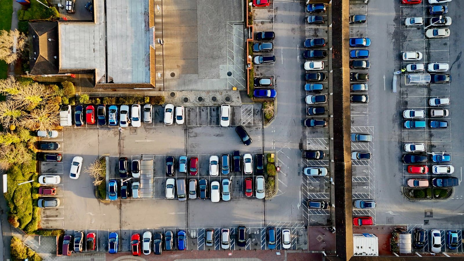 Vue aérienne d'un grand parking extérieur occupé par de nombreuses voitures, avec ses voies de circulation et ses bâtiments.